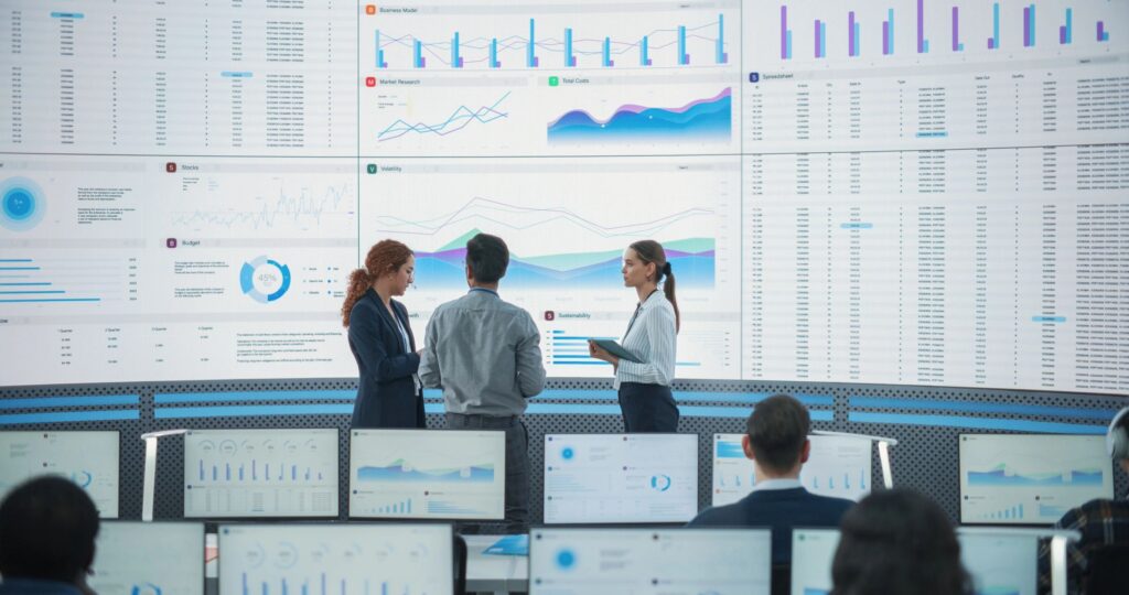 Wide Back Shot of Diverse Team of Data Scientists Discussing Graphs And Reports On Big Digital Screen In Monitoring Office. Multiethnic Employees Working On Desktop Computers For Deep Learning Company